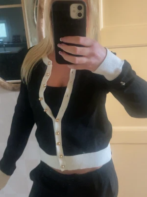 Svart kofta med vita detaljer och guldknappar - Snygg svart kofta med vita ribbade muddar och kant, dekorerad med guldfärgade knappar längs hela öppningen. Koftan har en croppad passform och är perfekt att styla över ett linne. Materialet ser ut att vara mjukt och stretchigt.