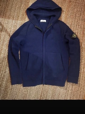 Mörkblå hoodie från Stone Island - Snygg mörkblå hoodie från Stone Island med dragkedja framtill och två stora fickor. Klassisk huva och ikonisk logopatch på vänster ärm. Tillverkad i ett mjukt och tjockt material som känns riktigt lyxigt. Perfekt för dig som gillar streetwear och vill ha något stilrent.