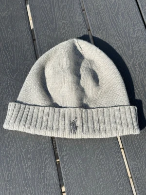 Ljusgrå stickad mössa Polo Ralph Lauren - Snygg ljusgrå stickad mössa från Polo Ralph Lauren med uppvikt kant och broderad logga framtill. Tillverkad i mjukt material som håller dig varm och passar perfekt till kalla dagar. Klassisk och stilren design som funkar till det mesta.