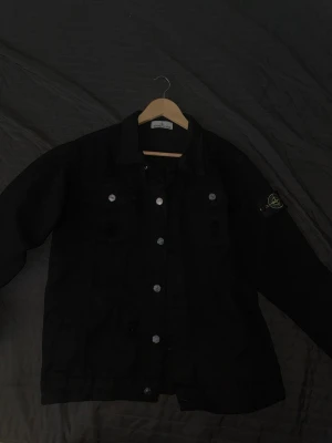 Svart jeansjacka från Stone Island - Snygg svart jeansjacka från Stone Island med klassisk krage, silverfärgade knappar och ikonisk logotyp-patch på ärmen. Jackan har två bröstfickor med knapp och raka ärmslut. Perfekt för dig som gillar streetwear och vill sticka ut.
