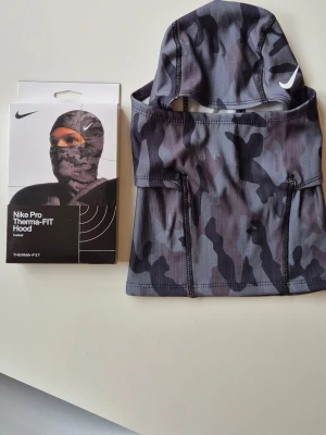 Nike Pro Therma-FIT camo huva - Nike Pro Therma-FIT huva i grått och svart camouflagemönster. Tillverkad i ett mjukt och stretchigt syntetmaterial som andas. Perfekt för kalla dagar när du vill hålla dig varm och skyddad. Snygg Nike-logga på sidan.