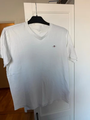 Vit v-ringad t-shirt från GANT - Vit t-shirt från GANT med v-ringning och diskret logga på bröstet. Klassisk passform och tillverkad i mjuk bomull, perfekt för en clean och stilren look. Passar dig som gillar enkelhet och kvalitet.