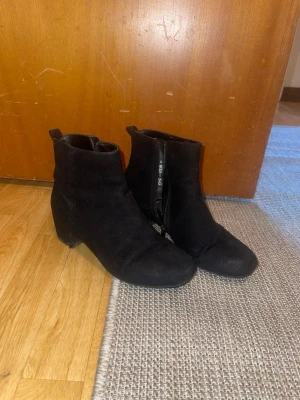 Svarta mockastövletter med klack - Säljer ett par svarta stövletter i mockaimitation med dragkedja på insidan och bred klack. Skorna har en stilren och enkel design som passar till många olika outfits. Perfekta för dig som vill ha ett par klassiska boots med lite höjd.