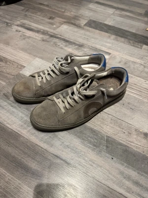 Oliver cabell skor - Snygga gråa sneakers i mocka med rundad tå och vita snören. Skorna har en blå detalj på hälen och en diskret cirkelformad design på sidan. Perfekta för dig som gillar en clean och stilren look. Finns tecken på användning och är bara smuts orkar själv inte tvätta dom vilket bör göra så dom ser nya ut säljer eftersom jag inte använder dom, har låda och är från deras orginal hemsida 