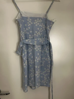 Ljusblå blommig sommarklänning - Söt ljusblå klänning med vita små blommor och smala axelband. Klänningen har en rak passform, avtagbart midjeband och är knälång. Perfekt för varma dagar och har en luftig känsla. Materialet ser ut att vara ett lätt tyg, troligen bomull eller viskos.