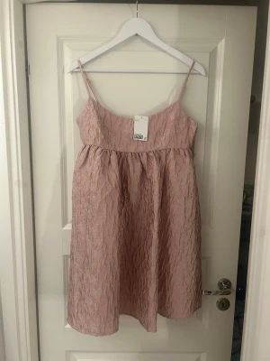 Ljusrosa klänning med struktur - Söt ljusrosa klänning med smala axelband och vacker struktur i tyget. Klänningen har en omlottliknande överdel och ett lätt utställt kjolparti. Perfekt för sommarens alla tillfällen och ger en romantisk vibe.