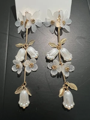 Långa örhängen med blommor och pärlor - Unika örhängen med hängande vita blommor och pärlliknande detaljer. Örhängena har guldfärgade stjälkar och blad, samt genomskinliga och vita blommor i plast eller akryl. Perfekta för att ge en romantisk touch till din stil.