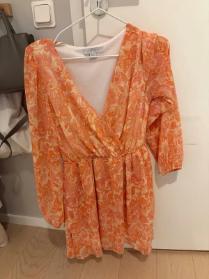 Orange mönstrad omlottklänning Amisu - Superfin kort omlottklänning från Amisu i orange och vitt med blommigt mönster. Klänningen har v-ringning, långa ärmar och resår i midjan. Perfekt för sommarens alla tillfällen!