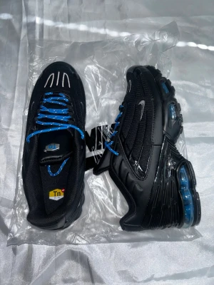 Nike Air Max Plus Tn Svart/Blå - Nike Air Max Plus Tn sneakers i svart med blå detaljer och blå snören. Skorna har synliga Air Max-bubblor i sulan, Tn-logga på hälen och mesh- och syntetmaterial på ovandelen. Swoosh-logga i silver på sidan. Riktigt snygga och sportiga sneakers.