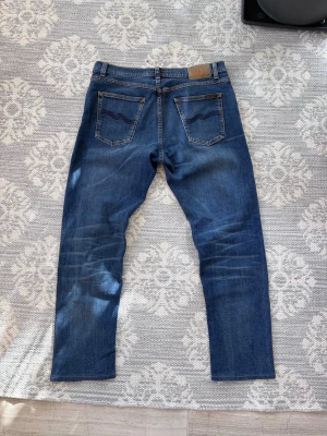 Blå jeans från Nudie Jeans - Säljer ett par klassiska blå jeans från Nudie Jeans med snygga slitningar och raka ben. Jeansen har fem fickor, kontrastsömmar och en läderpatch bak i midjan. Perfekta för en avslappnad och trendig stil. Lite förkortade