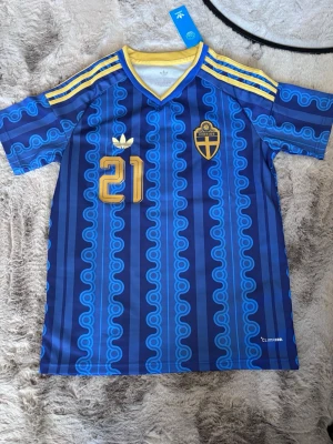 Sverige fotbollströja KULUSEVSKI #21 - Blå och gul Sverige fotbollströja från Adidas med KULUSEVSKI och nummer 21 på ryggen. Tröjan har retroinspirerat mönster, gula ränder på axlarna och broderat landslagsmärke. Tillverkad i lätt och ventilerande material, perfekt för match eller träning.