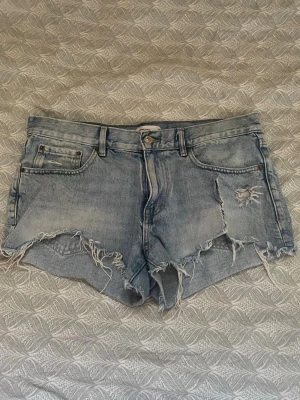 Ljusblå denimshorts med fransar - Säljer ett par ljusblå denimshorts med råa, fransiga kanter och slitna detaljer framtill. Klassisk femficksmodell med bälteshällor och knappstängning. Perfekta för sommardagar och en avslappnad look.