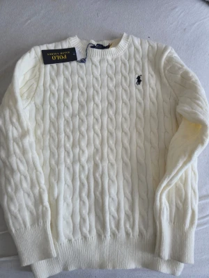 Vit kabelstickad tröja Polo Ralph Lauren - Kabelstickad vit tröja från Polo Ralph Lauren med rund halsringning och broderad logga på bröstet. Tröjan har långa ärmar och ribbade muddar vid ärmslut och nederkant. Perfekt för en stilren och klassisk look.Obs den har en liten hål kolla bild