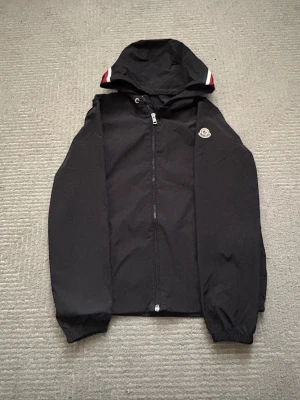 Svart Moncler vindjacka med huva - Snygg svart vindjacka från Moncler med dragkedja framtill och huva. Jackan har elastiska muddar vid ärmsluten och Moncler-logga på vänster ärm. Detaljer i vitt och rött på huvan ger en sportig touch. Perfekt för blåsiga dagar.