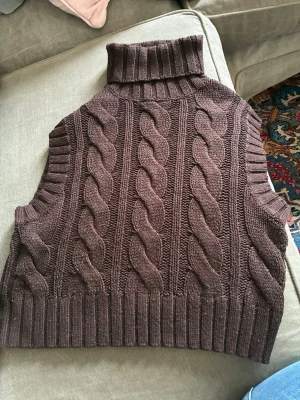 Brun kabelstickad väst med polokrage - Säljer en brun kabelstickad väst med hög polokrage och breda ribbade kanter. Västen är gjord i ett mjukt stickat material och har ett klassiskt flätmönster som ger en cool och tidlös look. Perfekt att styla över en långärmad tröja.