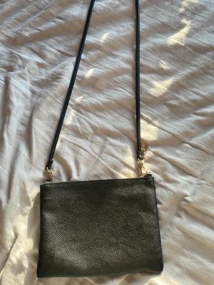 Svart axelväska från H&M - En stilren svart axelväska från H&M i slätt konstläder med guldfärgade detaljer och dragkedja. Väskan har en lång, smal axelrem och ett rymligt huvudfack med innerficka. Perfekt för att bära det viktigaste med enkel och snygg look.