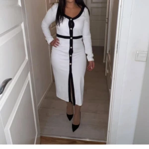 Vit långklänning med svarta detaljer - Stilren vit långklänning med svarta kanter och knappar framtill. Klänningen har lång ärm, v-ringning och markerad midja med svart bälteffekt. Perfekt för dig som gillar klassisk och elegant stil.