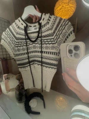 Stickad topp med mönster från Zara - Kortärmad stickad topp från Zara i beige och svart med grafiskt mönster. Tröjan har ribbad midja, snörning vid halsen och rundad halsringning. 