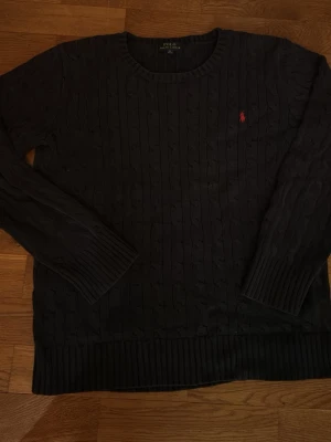 Svart kabelstickad tröja Polo Ralph Lauren - Kabelstickad svart tröja från Polo Ralph Lauren med rund halsringning och röd broderad logga på bröstet. Långa ärmar och ribbade muddar vid ärmslut och nederkant. Klassisk och stilren design som passar till mycket.