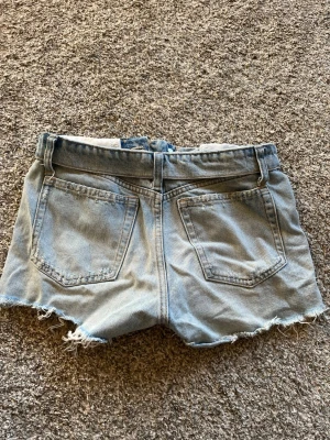 Ljusblåa jeansshorts med slitningar - Säljer ett par ljusblåa jeansshorts med fransiga kanter och slitna detaljer framtill. Perfekta för sommaren!