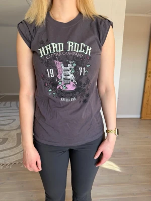 Hard Rock svart t-shirt med tryck - Cool grå t-shirt med stort tryck fram och bak. Motiv med elgitarr och text i vitt, rosa och mintgrönt. T-shirten har uppknäppta ärmar och har en avslappnad passform. Perfekt för dig som gillar musik och rockig stil. Använd en gång. 