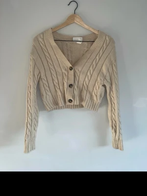 Beige kabelstickad kofta från Monki - Snygg beige kabelstickad kofta från Monki i croppad modell. Koftan har djup v-ringning, tre knappar framtill och långa ärmar. Perfekt att slänga över en topp för en trendig lager-på-lager-look.