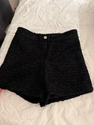 Svarta fluffiga högmidjade shorts i boucle - Säljer ett par svart högmidjade shorts i bouclé-look med knapp och dragkedja framtill (glittrar lite) Shortsen har en mjuk, texturerad yta och sitter snyggt på kroppen. Perfekta för sommarens alla tillfällen. Storlek M men skulle säga mer som xs-S 