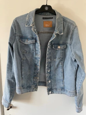 Ljusblå jeansjacka från ONLY - Snygg ljusblå jeansjacka från ONLY med klassisk krage, två bröstfickor med lock och silverfärgade knappar. Jackan har en rak passform och är tillverkad i mjukt denimtyg. Perfekt att slänga över en hoodie eller t-shirt.