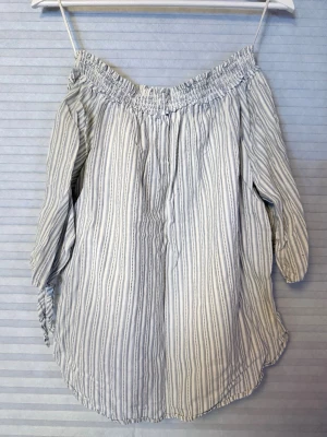 Randig offshoulder blus - Snygg offshoulder blus med vita och blå smala ränder. Blusen har resår upptill och vida ärmar som ger en avslappnad känsla. Tillverkad i ett lätt och luftigt tyg, perfekt för varma dagar.