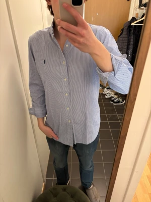 Blårandig skjorta från Ralph Lauren - Klassisk blårandig skjorta från Ralph Lauren med button-down krage och broderad logga på bröstet. Skjortan har långa ärmar, vita knappar och är tillverkad i bomull. Perfekt till jeans eller chinos för en clean och stilren look.