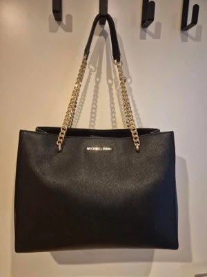 Svart Michael Kors handväska i läder - Stilren svart handväska från Michael Kors i strukturerat läder med guldfärgade kedjehandtag och logga framtill. Väskan har en fyrkantig form och rymligt huvudfack, perfekt för att bära med sig allt man behöver. Klassisk och trendig design med exklusiva detaljer.