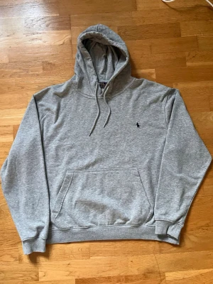 Grå hoodie från Polo Ralph Lauren - Klassisk grå hoodie från Polo Ralph Lauren med broderad marinblå logga på bröstet. Hoodien har huva med dragsko, stor magficka och ribbade muddar vid ärmslut och nederkant. Tillverkad i mjuk bomullsblandning för maximal komfort.