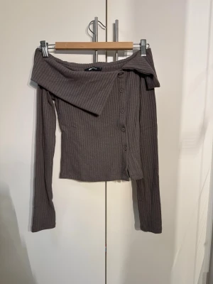 Brun ribbad off shoulder topp Gina Tricot - Trendig brun ribbad off shoulder topp från Gina Tricot. Toppen har långa ärmar, knappar framtill och en bred krage som ger en snygg asymmetrisk look. Perfekt för dig som vill ha en unik och stilren topp till garderoben.