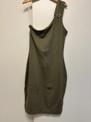 Olivgrön oneshoulder klänning (kort) - Snygg olivgrön klänning i oneshoulder-modell med dekorativt spänne på axeln. Klänningen är figurnära och kort, perfekt för dig som vill ha en stilren och trendig look. Endast provad, aldrig använd, legat i garderob i några år nu. 