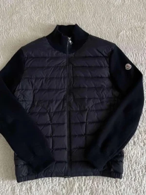 Moncler Cardigan - Stilren svart pufferjacka från Moncler med quiltad front och ärmar i mjukt tyg. Jackan har hög krage, hel dragkedja och Moncler-logga på ena ärmen. Perfekt för kalla dagar och ger en clean look.