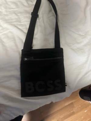 Svart axelväska från BOSS - Snygg svart axelväska från BOSS med en mer slim look till den, den är perfekt för dem som inte gillar stora axelremsväskor. Den är i bra skick och är 6 månader gammal där jag inte använde den så mycket vid vintern. Dragkedjan har tappat delen som man drar fast man kan köpa delen och fixa det. Kan pruta ner produkten.
