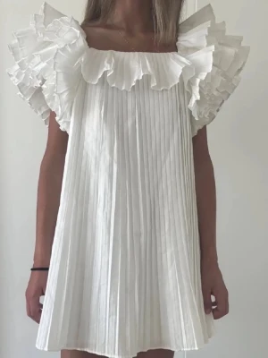 Vit plisserad klänning med volangärmar - Superfin vit kortklänning med plisserat tyg och stora volangärmar. Klänningen har en luftig passform och är perfekt för sommarens alla tillfällen. Den är ärmlös och har en söt, romantisk look med mycket volym upptill.