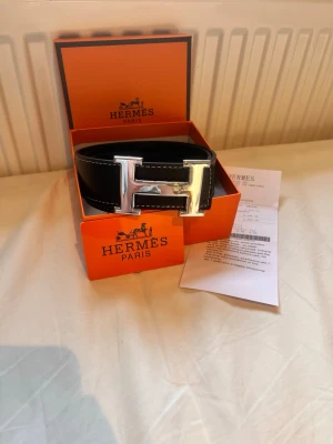 Svart skärp med H-spänne från Hrmès - Stilrent svart skärp från H med ikoniskt H-formad spänne i silver. Skärpet är tillverkat i exklusivt skinn och levereras i original orange box med tillhörande certifikat. Perfekt för att lyfta din outfit med en lyxig touch.