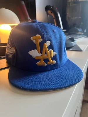 Blå LA Dodgers keps från New Era - Snygg blå keps från New Era med LA Dodgers-logga i gult och vitt framtill, dekorerad med molndetaljer. Klassisk flat brim och broderade detaljer på sidan. Perfekt för dig som vill sticka ut med en unik och trendig accessoar. Hatstorlek 7 3/8 eller 58,7 cm. 