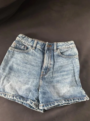 Blå jeansshorts från Lindex - Klassiska blå jeansshorts från Lindex med fem fickor och knappgylf. Shortsen har en avslappnad passform och är perfekta för varma dagar. Snyggt slitna detaljer och tidlös design som funkar till allt.