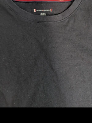 Svart t-shirt från Tommy Hilfiger - Klassisk svart t-shirt från Tommy Hilfiger med rund hals och diskret logga på bröstet. Tillverkad i mjuk bomull och har en normal passform. Perfekt basplagg för alla tillfällen.