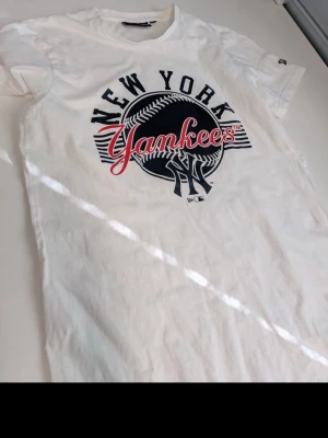 Vit New York Yankees t-shirt - Snygg vit t-shirt från New Era med stort New York Yankees-tryck i svart och rött framtill. Klassisk rund hals och korta ärmar. Perfekt för dig som gillar sportig streetstyle och vill visa vilket lag du hejar på.
