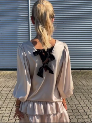 Neo Noir set med volanger och rosetter strl S - Säljer ett superfint tvådelat set i beige med puffärmar och volanger. Toppen har öppning i ryggen med två stora svarta rosetter som detalj. Kjolen är kort med flera lager volanger och smock i midjan. Perfekt för dig som gillar romantisk och trendig stil.