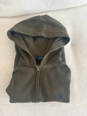 Olivgrön hoodie från Polo Ralph Lauren - Snygg olivgrön hoodie med dragkedja från Polo Ralph Lauren. Klassisk design med huva, kängurufickor och blå broderad logga på bröstet. Skick: 8,5/10😮‍💨🔥👍🏻. Storlek: 152-164😮‍💨🔥👍🏻🍾. Perfekt för en avslappnad och grisch stil. 🤙🏼🤙🏼🤙🏼