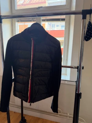 Moncler cardigan - Snygg svart pufferjacka från Moncler med quiltad front och stickade ärmar. Dragkedjan har röd, vit och blå detalj och jackan har en hög krage. Moncler-logga på ärmen. Perfekt för kalla vinterdagar och riktigt trendig look.