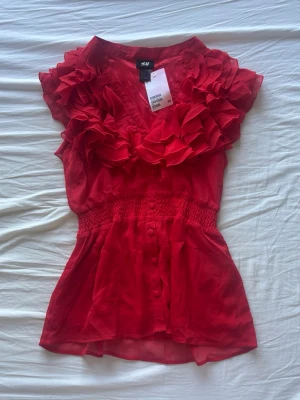 Röd volangblus från H&M - Snygg röd blus från H&M med volanger längs v-ringningen och axlarna. Blusen har knappar framtill, markerad midja med smock 