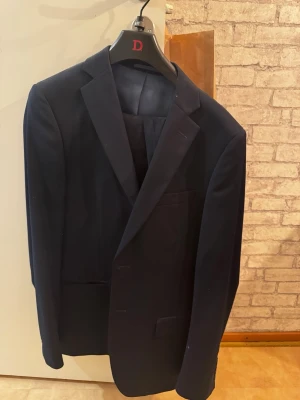 Mörkblå klassisk kavaj - Snygg mörkblå kostym (kavaj och kostymbyxor) i klassisk modell med enkelknäppning. Passar både till kostymbyxor som ingår med och jeans för en stilren look. Endast använd en gång och i nyskick – ren, fräsch och utan skador. Två framfickor och en bröstficka.  Storlek L för både kostym och byxor
