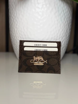 Brun korthållare från Coach - Snygg brun korthållare från Coach med klassiskt logomönster och guldfärgad metallogo på framsidan. Praktisk och stilren design med flera fack för kort. Perfekt accessoar för dig som vill ha koll på korten utan att ta plats.