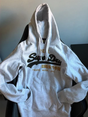 Ljusgrå Superdry hoodie - Säljer denna ljusgrå hoodien från superdry! Den har tjocka snören som är skitsnygga och en speciell modell med konfettiliknande tema på ”superdry” texten. Den är i S för herr och M för kvinna, men det är bara att skriva vid andra funderingar osv. Den är i så fint skick du kan hitta utan minsta skråma. 