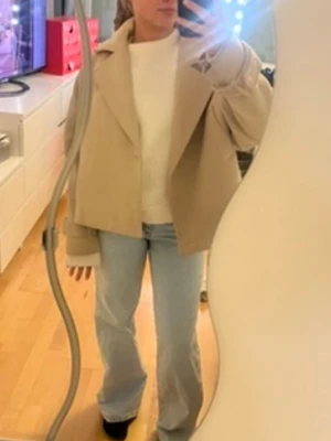 Beige kort kappa från Nelly - Snygg beige jacka från Nelly med bred krage och croppad passform. Jackan har långa ärmar med spänndetaljer vid ärmsluten och en stilren design. De tillkommer även ett midjeband som jag kan skicka med men som jag personligen aldrig använt då man även kan stänga jackan med knappar.